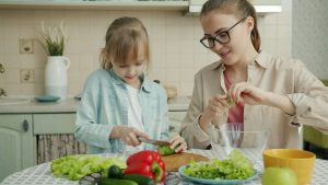 Adorable little girl is helping mother in kitchen cutting vegetables for salad and talking making dinner together. Healthy nutrition, lifestyle and people concept.
